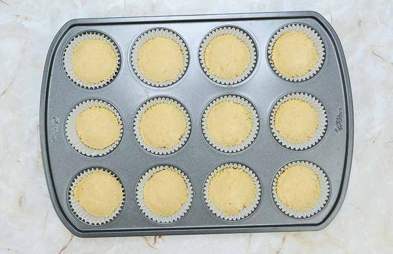 A muffin tin containing twelve paper-lined cups filled with uncooked candy corn cheesecake batter, ready for baking. The tin is placed on a light-colored countertop.