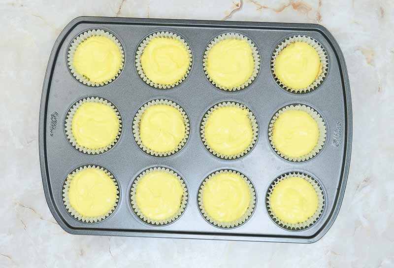 A muffin tin with twelve slots, each filled with a light yellow candy corn cheesecake batter in paper liners, is placed on a marble countertop. The batter appears smooth and evenly distributed in each liner, ready to be baked.