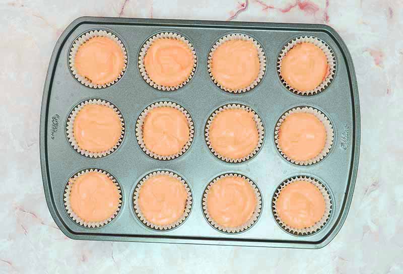 A muffin tin filled with 12 unbaked cupcakes in white paper liners, their light orange batter evoking the hues of candy corn cheesecake. The tin rests on a pinkish marble countertop.