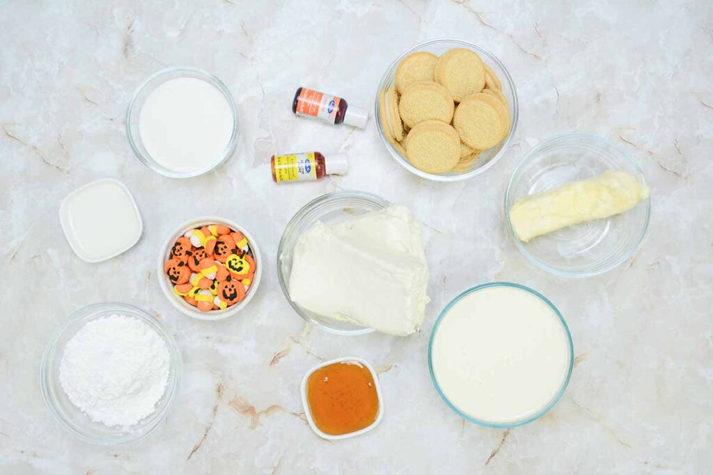 A top-down view of baking ingredients on a marble surface reveals the essentials for a delightful candy corn cheesecake. Items include cream cheese, butter, vanilla extract, food coloring, cookies, powdered sugar, milk, candy corn, decorative candy pumpkins, and a small dish of caramel sauce.
