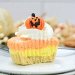 A festive dessert featuring a layered mini candy corn cheesecake with cream-colored, orange, and yellow sections, topped with whipped cream and a small pumpkin decoration. The treat sits on a plate with an autumn-themed background.