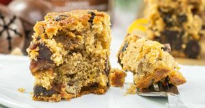 A close-up of a slice of homemade fruit cake on a white plate showcases its moist texture, filled with various nuts and dried fruits. In the background, another slice of these delectable fruitcake bites is partially visible with a fork next to it. The background is softly blurred.