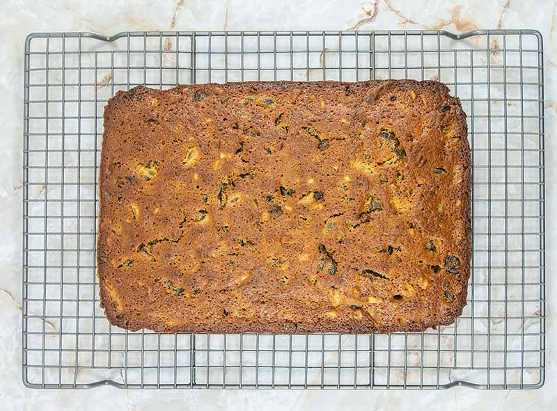 A freshly baked, rectangular loaf of zucchini bread resting on a cooling rack. The bread, reminiscent of fruitcake bites, has a golden-brown crust with visible specks of green zucchini and small pieces of nuts embedded throughout. The cooling rack and bread are placed on a marble surface.