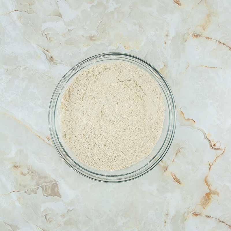 A clear glass bowl filled with a fine beige powder, placed on a marble surface with light brown and tan veining. The image is taken from directly above, showcasing the even texture of the powder inside the bowl, reminiscent of the delicate ingredients used in making fruitcake bites.