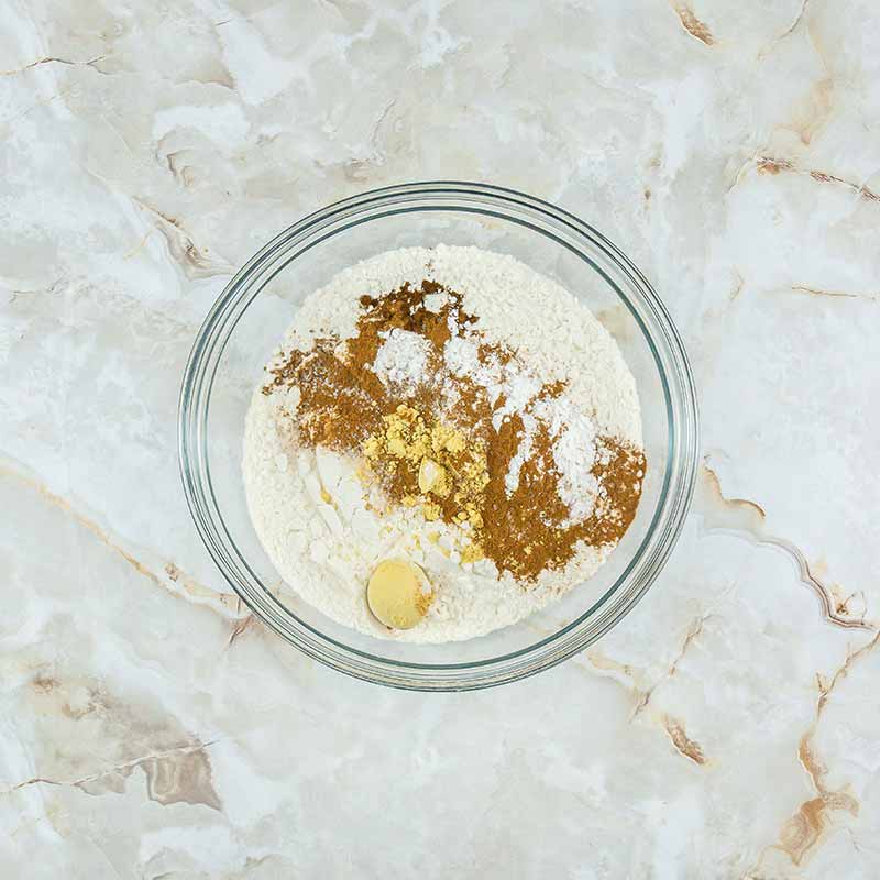 A clear glass bowl containing flour, brown sugar, white sugar, cinnamon, and other spices rests on a marbled countertop, poised to create delectable fruitcake bites. The ingredients are not yet mixed, displaying distinct layers and colors.