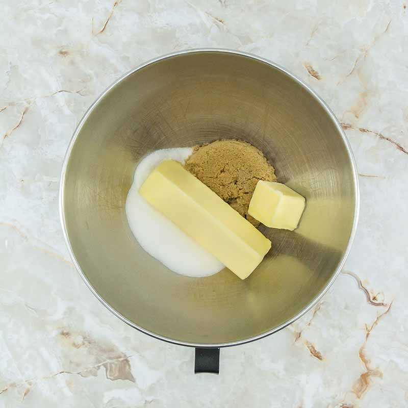 A stainless steel mixing bowl containing a stick of butter, a small additional piece of butter, brown sugar, and white sugar, perfect for preparing fruitcake bites, is placed on a marbled countertop.