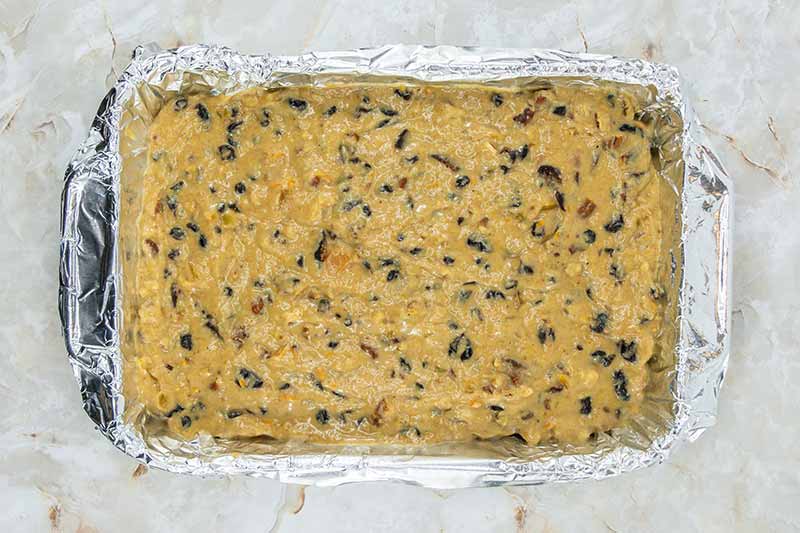 A rectangular baking dish lined with aluminum foil is filled with raw, light brown cake batter mixed with dark chocolate chips and nuts. These fruitcake bites are then placed on a marbled countertop, ready for the oven.
