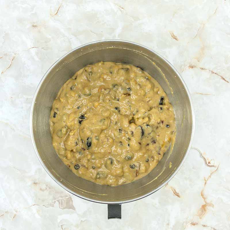 A stainless steel bowl filled with cookie dough mixed with chocolate chips and bits of fruitcake bites, set on a light-colored marble countertop. The dough appears to be well-blended and ready for baking.