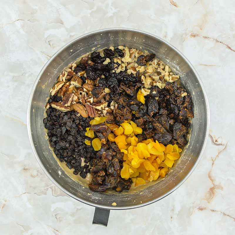 A metal mixing bowl filled with various chopped dried fruits and nuts, including dried apricots, raisins, walnuts, and pecans, evokes the rich flavors of fruitcake bites against a light marbled background.