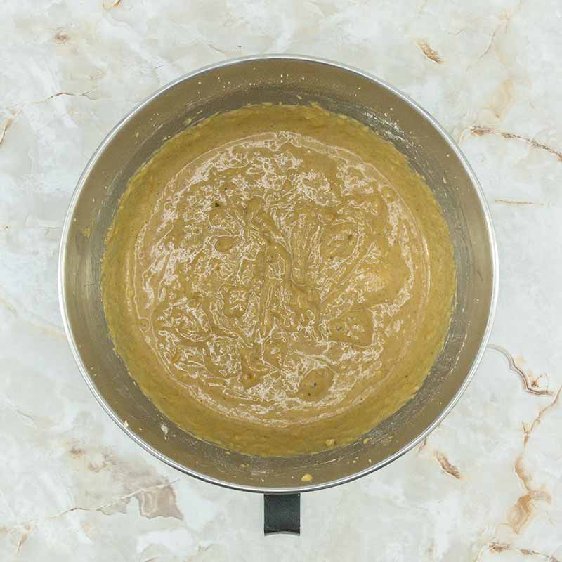 A large metal mixing bowl filled with thick, light brown batter rests on a marble countertop. The batter, likely destined for fruitcake bites, appears to have been recently mixed, with swirled patterns visible on the surface.