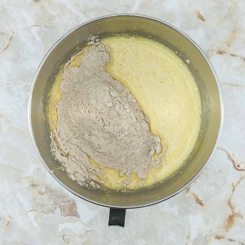 A metal mixing bowl filled with yellow batter and a partially mixed portion of dry, light brown ingredients sits on a marbled countertop. Intended for fruitcake bites, the dry ingredients are heaped on one side, clearly not yet fully incorporated into the batter.