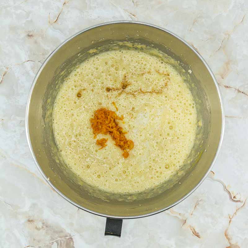 A stainless steel mixing bowl contains a yellowish batter with small clumps of orange zest and a dash of cinnamon on top, ready to be stirred for perfect fruitcake bites. The bowl rests on a marbled counter surface.