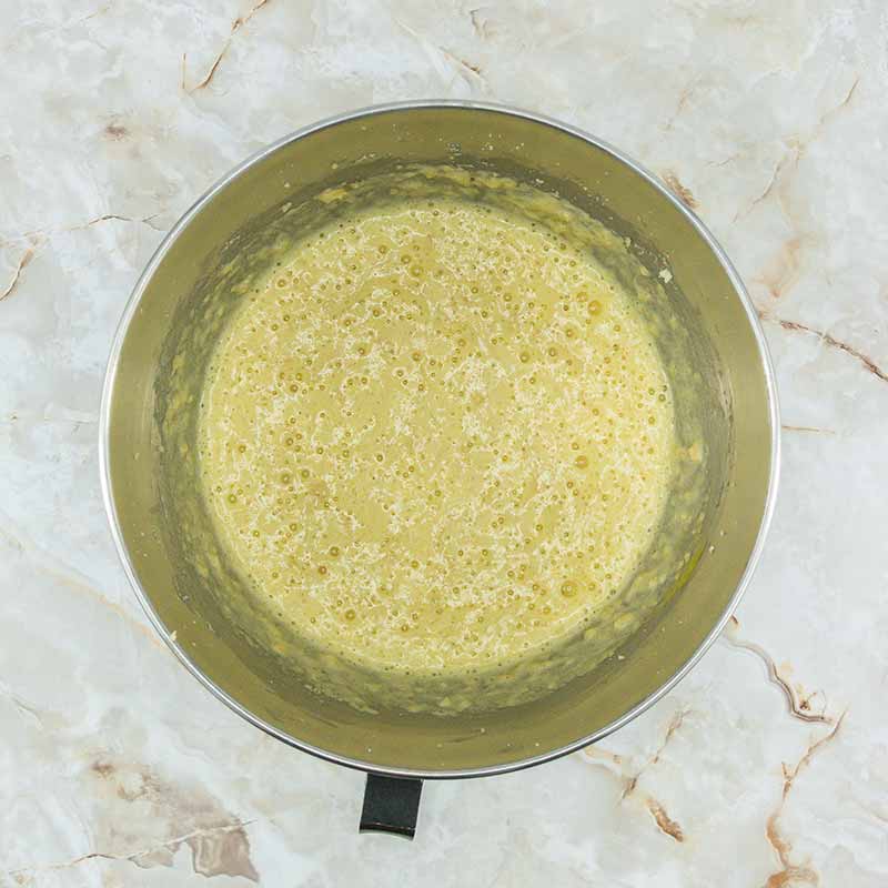 A metal mixing bowl filled with a frothy, pale yellow batter sits on a marble countertop. The batter, destined for delectable fruitcake bites, is smooth with small bubbles visible on the surface, indicating it has been well mixed or is in the process of reacting.