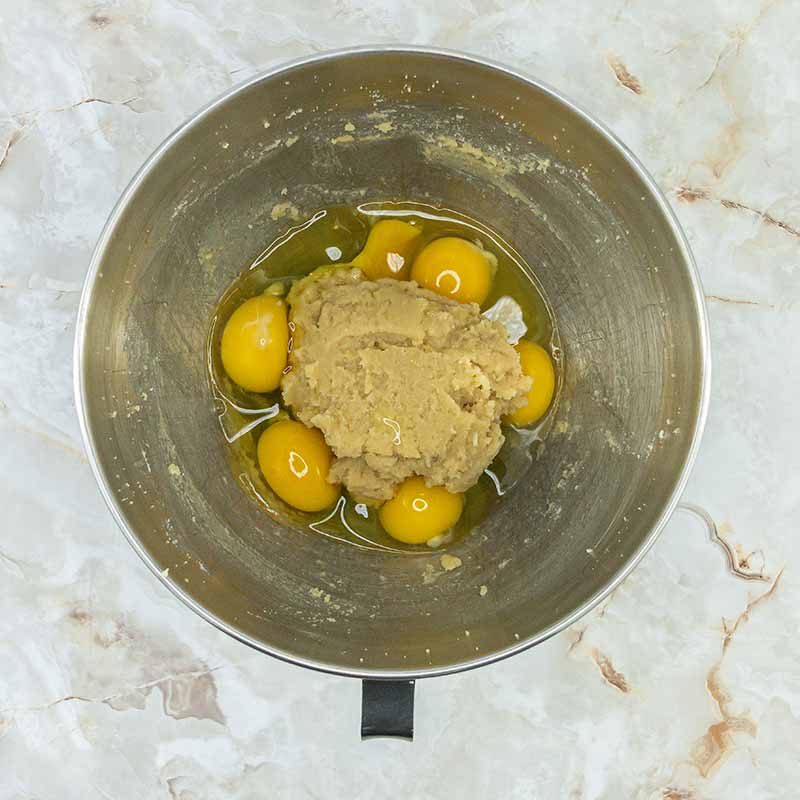 A metal mixing bowl containing cracked raw eggs and a dollop of brown sugar sits on a marble countertop, ready to be mixed into delicious fruitcake bites.