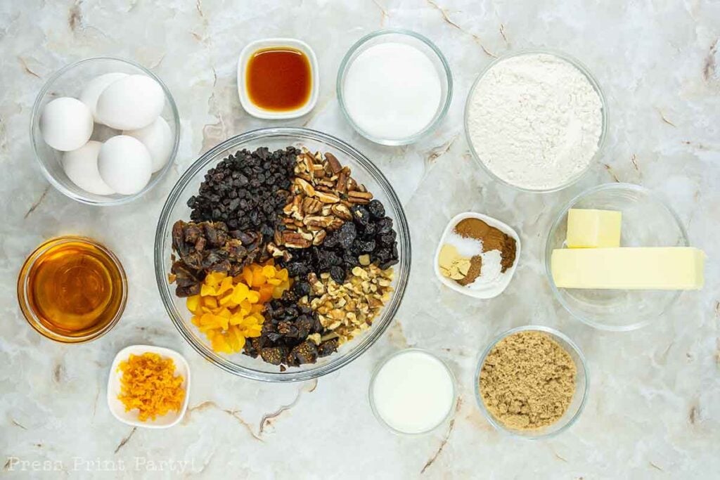 A top-view photo of several baking ingredients arranged on a marble surface, perfect for whipping up delicious fruitcake bites. Ingredients include bowls of eggs, vanilla extract, granulated sugar, flour, dried fruits, nuts, brown sugar, butter, orange zest, assorted spices, molasses, and milk.