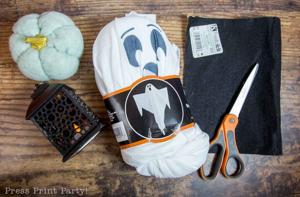 A table holds materials for a Halloween craft project: a soft fabric pumpkin, a black lantern with a candle inside, a white fabric with a ghost face design, a piece of black felt, a pair of scissors, and a felt piece with a price tag. "Press Print Party!" is in the corner.