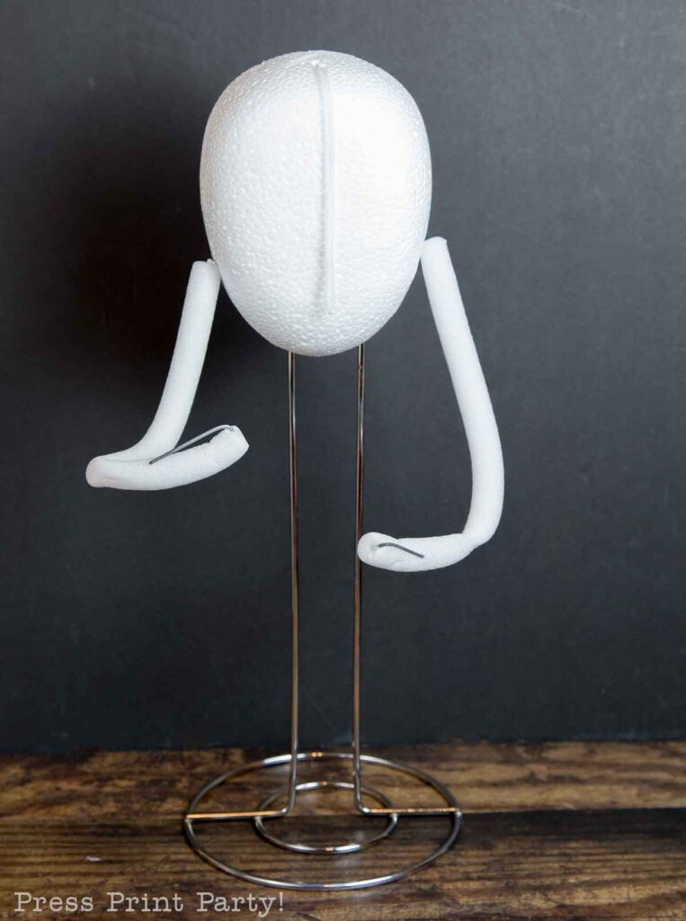 A simple paper towel stand with a white foam head and foam-padded wire arms displayed on a wooden surface against a dark background. The writing "Press Print Party!" is lightly visible at the bottom left corner.