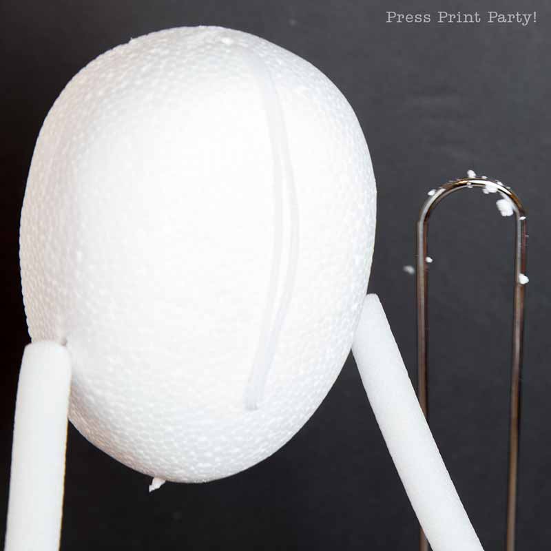 A close-up of a white Styrofoam egg with a seam running vertically. Two white pipe cleaners are attached on either side, possibly forming arms or legs. The background is black, with a metal clip visible to the right.