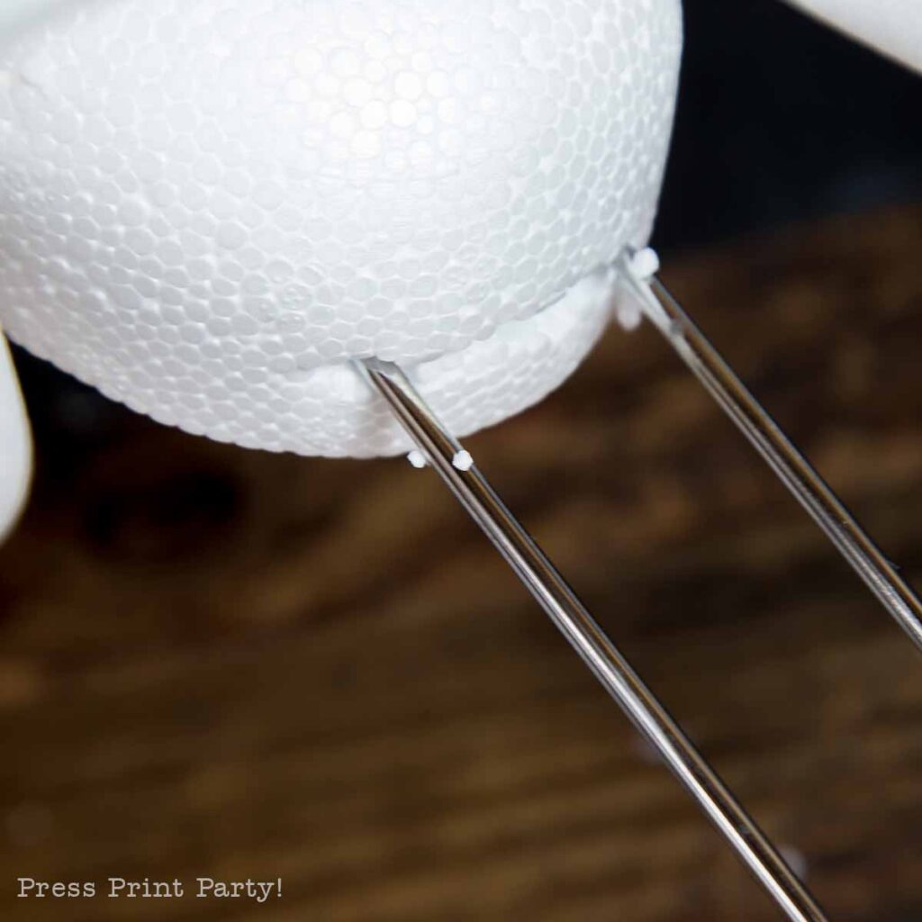 Close-up of a white Styrofoam sphere with metal towel bar running through it. The background is blurred, showing a dark wooden surface. The bottom corner has partially visible text, "Press Print Party!.