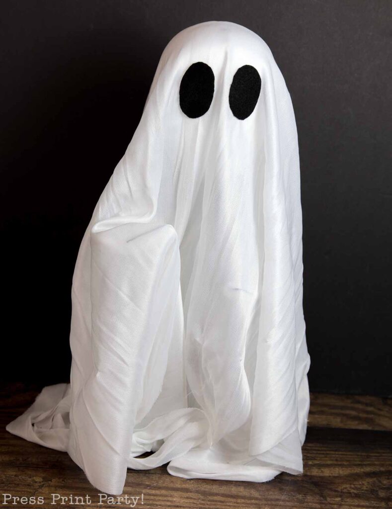 A Halloween ghost decoration featuring a white, flowing sheet with two large, oval black eyes. The ghost appears against a dark background, creating a spooky yet playful atmosphere.