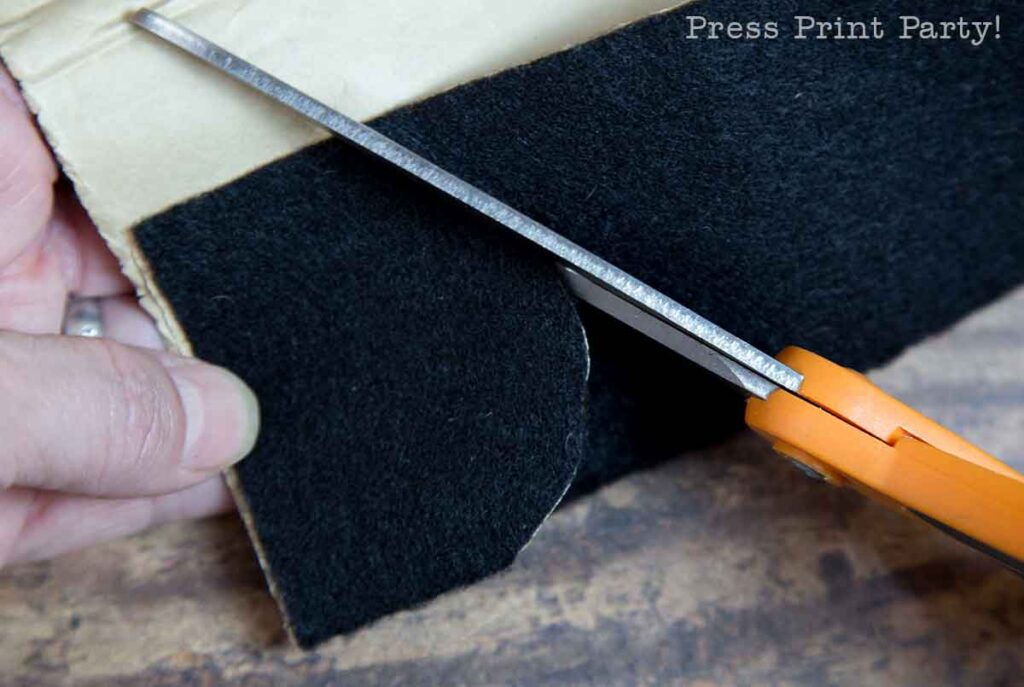 A hand holding orange-handled scissors is seen cutting through black felt fabric and brown paper. The background has a wooden texture. Text in the upper right corner reads "Press Print Party!.