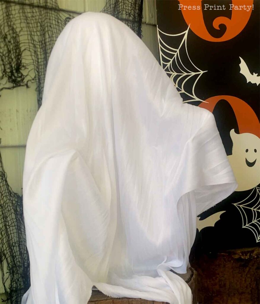 A person draped in a white sheet resembles a ghost, sitting with an eerie backdrop decorated with black spider webs, bats, and a smiling ghost. The scene evokes a Halloween theme.