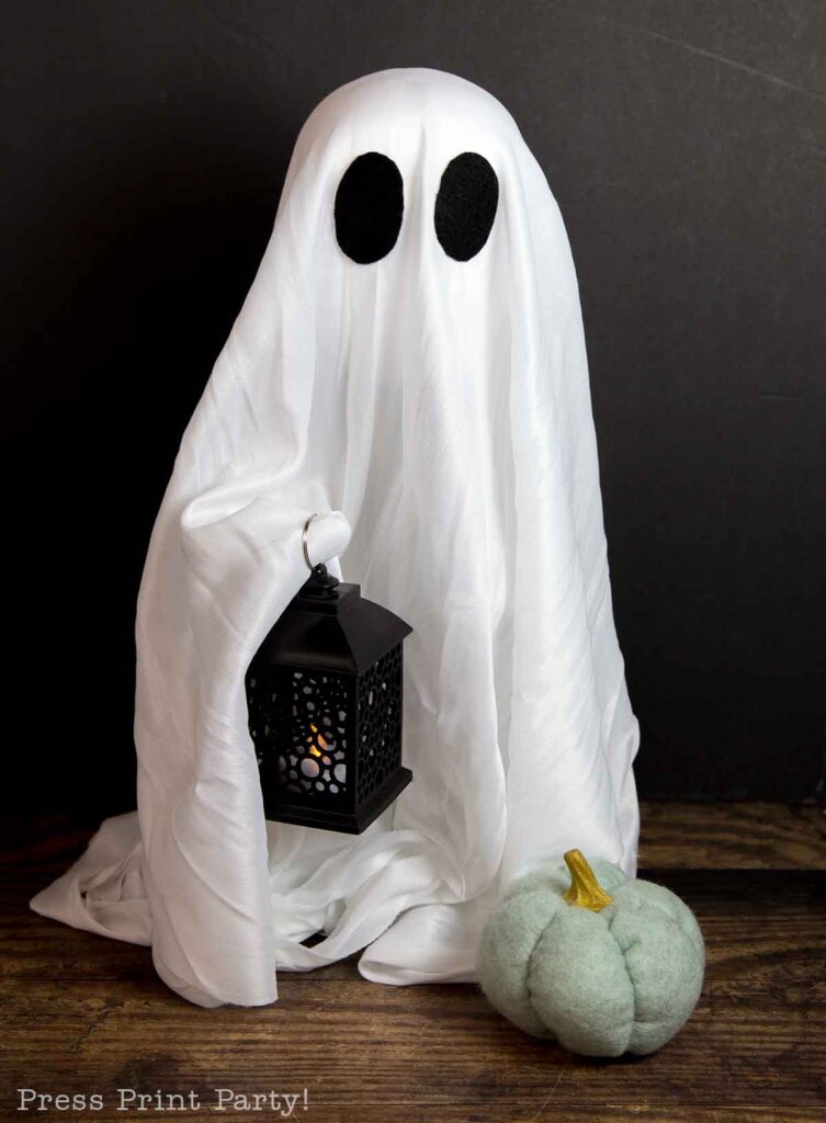 A ghost costume made of white fabric with black oval eyes, holding a black lantern with a small light. To the right of the ghost is a small, decorative green pumpkin. The background is dark, with a wooden surface underneath the costume and props.