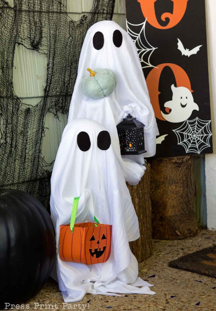 Two ghost decorations, draped in white cloth with black eye holes, stand with Halloween accessories. One holds a black lantern and the other a candy bucket shaped like a pumpkin. Cobweb decor, a pumpkin, and Halloween-themed props adorn the background.