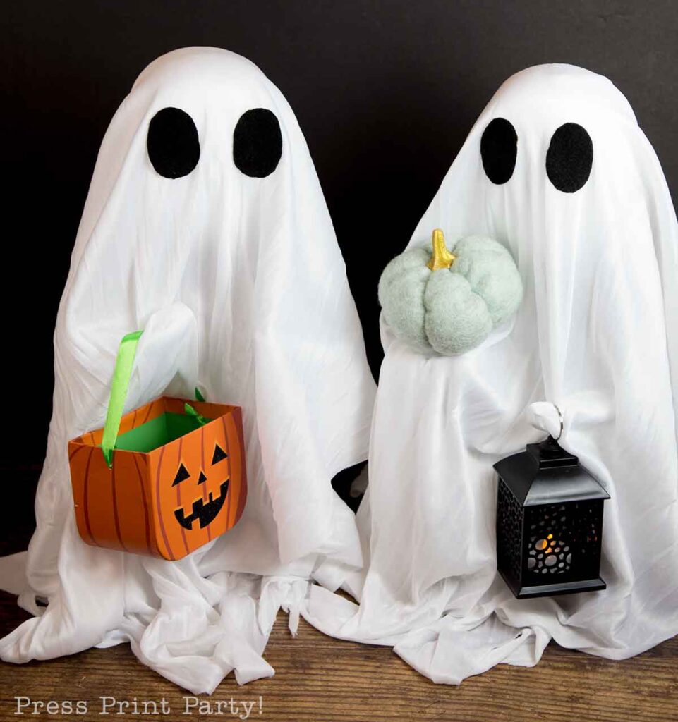 Two individuals dressed as ghosts with white sheets and black eye cutouts. One ghost holds a green-handled, pumpkin-shaped candy basket, and the other holds a lantern and a plush pumpkin. A textured floor and black backdrop complete the Halloween-themed setup.
