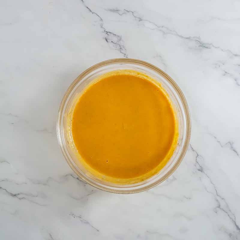 A clear glass bowl filled with smooth, orange-colored soup sits on a white marble surface, evoking the rich hues of a pumpkin pie. The creamy-textured soup has an even consistency, perfectly complemented by the subtle grey veining in the background.