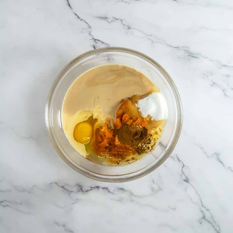 A clear glass bowl contains various ingredients including a cracked egg, sugar, spices, and a brown mixture for pumpkin pie cookies set on a white marble countertop.