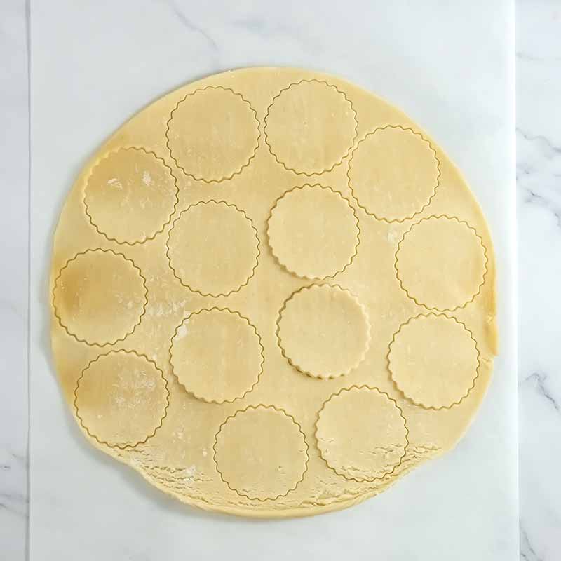 A round sheet of rolled-out dough on a marble surface, with multiple scalloped-edged circles cut out from it, ready for baking pumpkin pie cookies or filling.