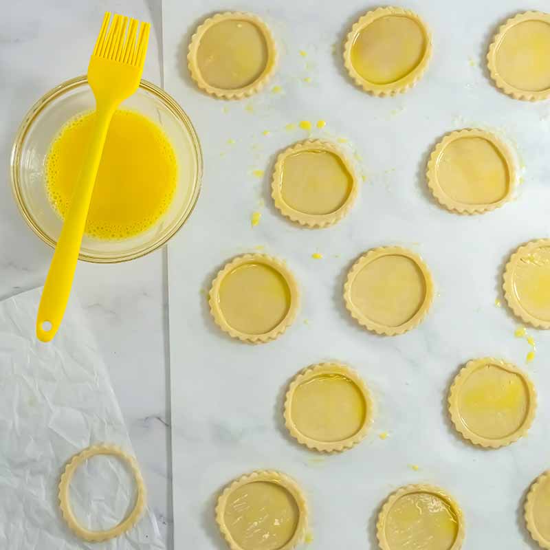 A baking sheet covered in parchment paper holds multiple uncooked pumpkin pie cookies with slightly raised edges. A small glass bowl filled with egg wash, and a yellow pastry brush, rest to the side. An extra pastry circle sits on the parchment paper.