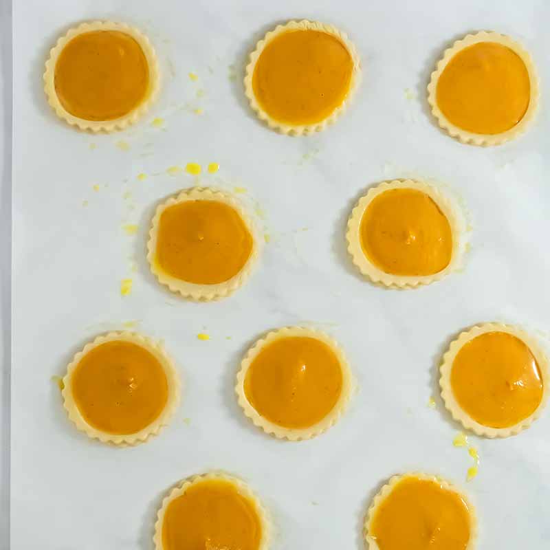 A baking tray with twelve unbaked pumpkin pie tarts arranged in a 3x4 grid. The tarts have fluted edges and are filled with smooth, orange pumpkin filling, reminiscent of a delightful pumpkin pie cookie, ready to be baked. The tray is lined with parchment paper.