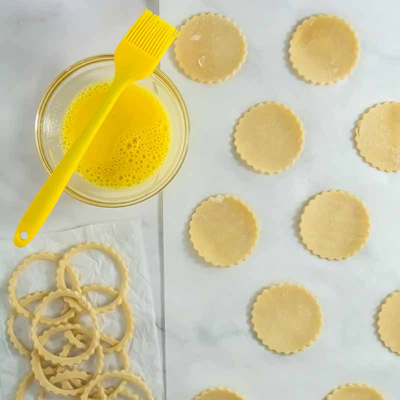A flat surface is covered with circular dough cutouts, ready to be transformed into pumpkin pie cookies. Next to them is a small bowl filled with yellow egg wash, and a yellow pastry brush rests on the bowl's rim. A separate pile of dough scraps lies off to the side.
