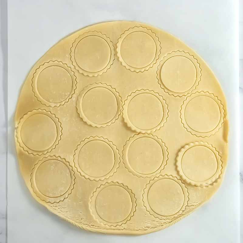 A circular sheet of dough with evenly spaced out scalloped-edge circles cut out of it, resting on a white surface. The dough has a smooth texture and the cut-out circles are ready to be turned into delightful pumpkin pie cookies for baking or cooking.