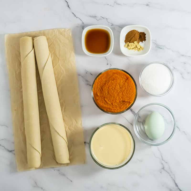 Ingredients for pumpkin pie on a marble countertop: two rolled pastry doughs, a small bowl of vanilla extract, a small bowl of spices, a bowl of sugar, a bowl of pumpkin puree, a bowl of sweetened condensed milk, and an egg. Imagine turning these staples into delightful pumpkin pie cookies!