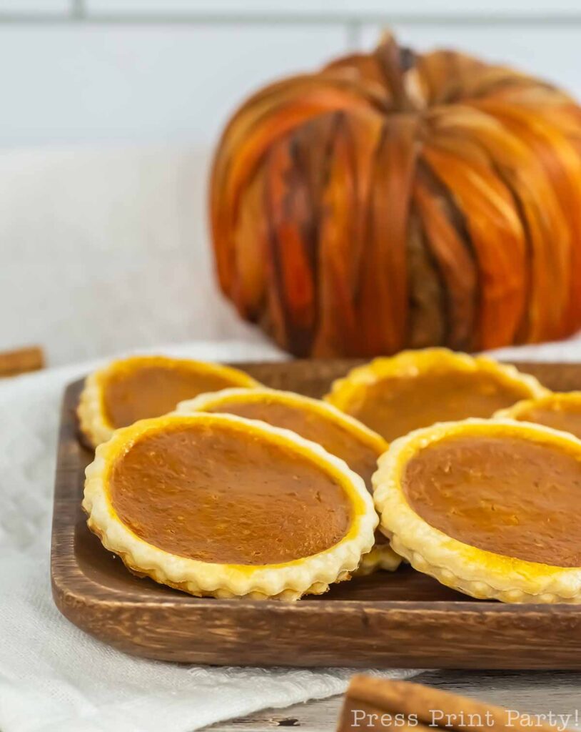 Mini pumpkin pies arranged on a wooden serving tray with a decorative pumpkin in the background. The pies have golden crusts and rich, pumpkin-colored filling. A white linen cloth is beneath the tray, making the cozy display perfect for any autumn gathering, like savoring a pumpkin pie cookie.