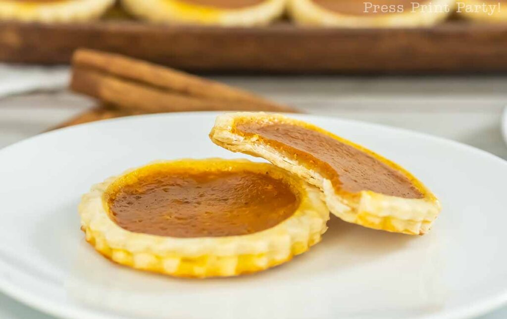 Close-up of two mini pumpkin pies on a white plate, resembling adorable pumpkin pie cookies. One pie is whole, while the other has a bite taken out of it, revealing the smooth pumpkin filling and golden crust. The background includes more pies and a couple of cinnamon sticks for added charm.