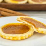Close-up of two mini pumpkin pies on a white plate, resembling adorable pumpkin pie cookies. One pie is whole, while the other has a bite taken out of it, revealing the smooth pumpkin filling and golden crust. The background includes more pies and a couple of cinnamon sticks for added charm.