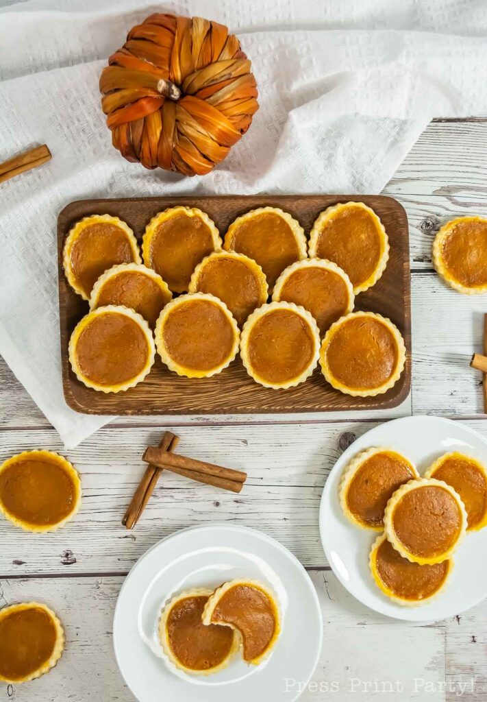 A display of mini pumpkin pies and pumpkin pie cookies is arranged on a wooden tray and two white plates, set on a rustic white table. Decorative cinnamon sticks and an ornamental pumpkin are placed nearby, adding to the autumnal theme.