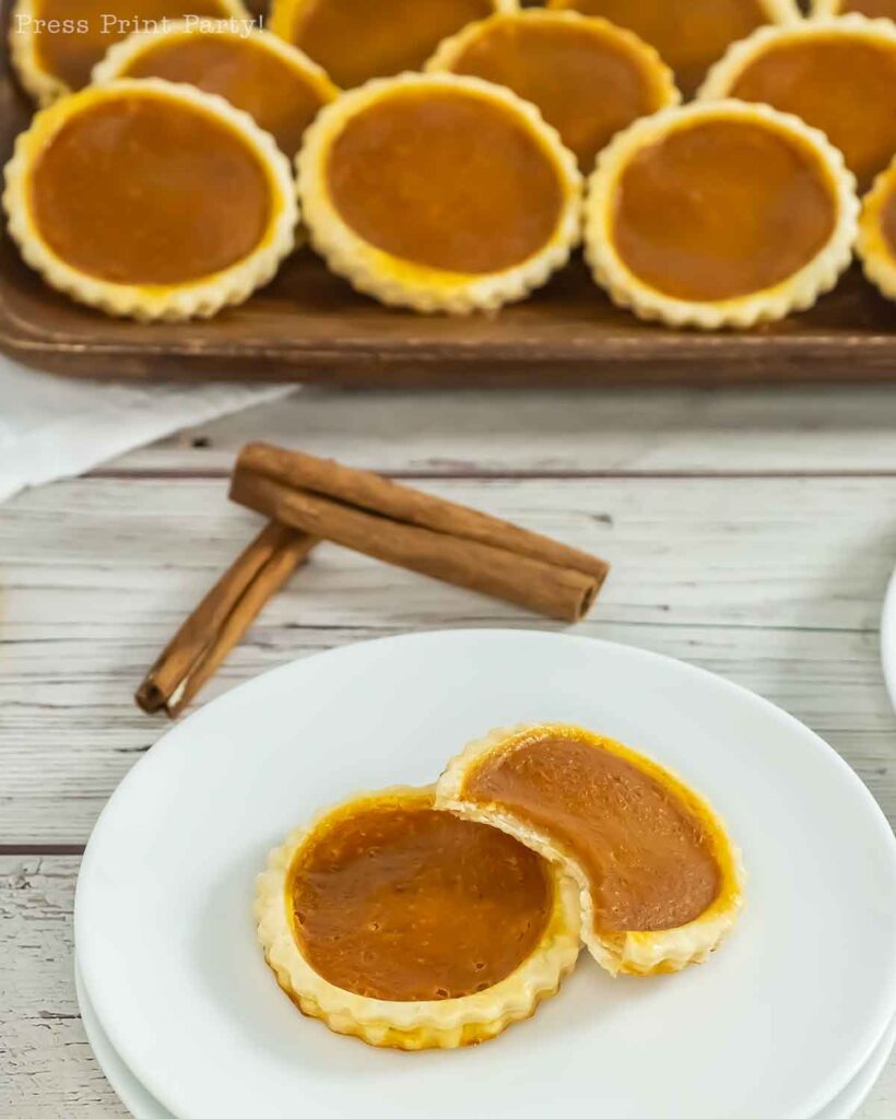 A plate with a partially eaten custard tart sits in the foreground, two cinnamon sticks artfully placed nearby. In the background, a tray filled with more custard tarts and pumpkin pie cookies is visible on the wooden table.