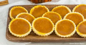 A wooden platter holds ten small, round pumpkin pie cookies with golden-brown crusts and a rich, orange filling. The platter is set on a white, textured surface, with a small portion of a cinnamon stick visible in the background.