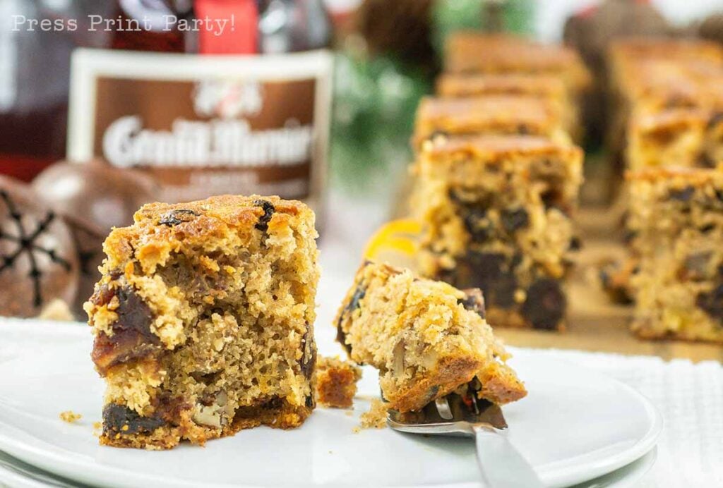A close-up of a slice of fruitcake on a white plate, with a fork beside it. The cake features visible nuts and dried fruits. In the background, there are more fruitcake bites, a blurred bottle labeled "Grand Marnier," and festive decorations.