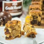 A close-up of a slice of fruitcake bites with nuts and dried fruits, partially eaten, placed on a white plate with a fork. In the background, there are more pieces of fruitcake and a bottle of Grand Marnier in soft focus. A holiday bell decoration is also visible.