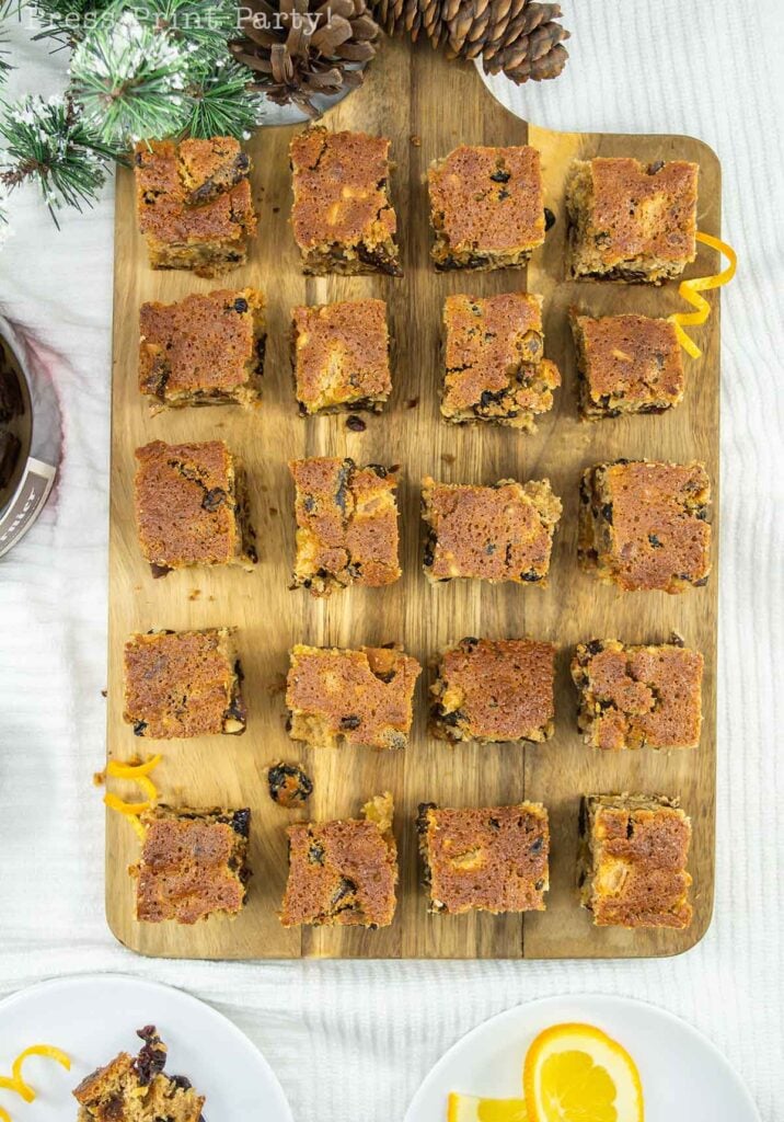 A wooden board holds twenty square fruitcake bites, arranged in a grid. Pinecones and evergreen branches decorate the top edge of the board. Orange garnishes and a slice of fruitcake are visible on a white plate in the bottom left corner.