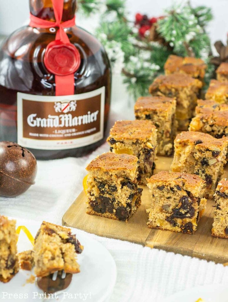 Squares of fruitcake bites are neatly arranged on a wooden board next to a bottle of Grand Marnier liqueur. Christmas decorations, including green fir branches and bells, are in the background, creating a festive holiday setting.