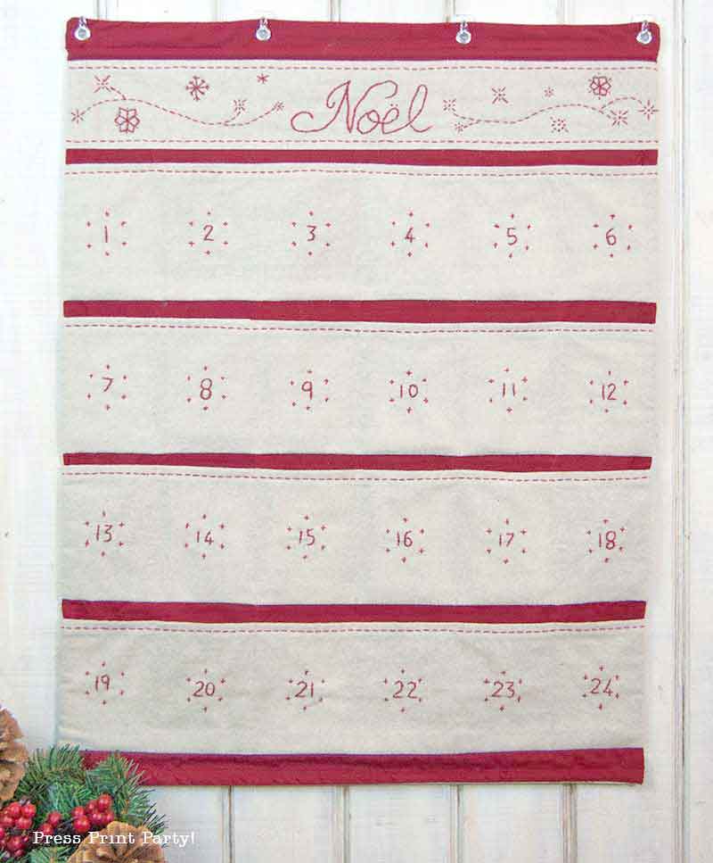 Fabric advent calendar with the word "Noël" embroidered at the top. It features 24 numbered pockets in rows, in red and cream colors, hanging against a wooden wall. A festive plant is in the bottom left corner.