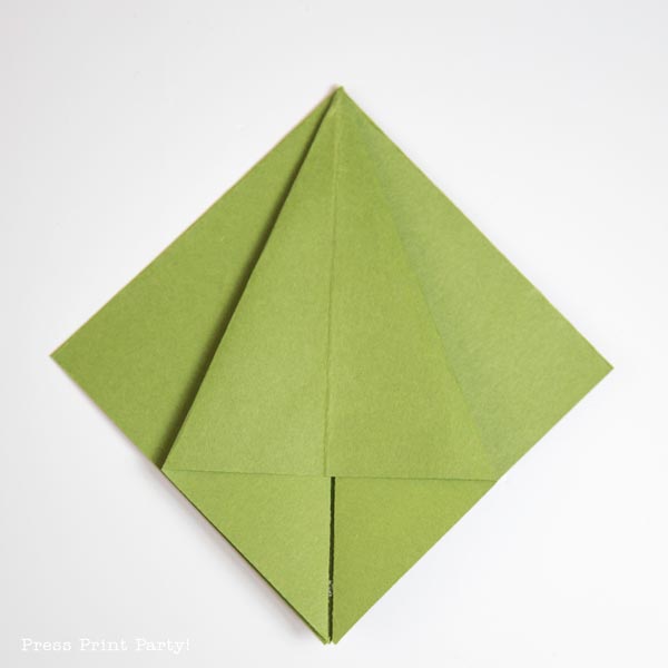 A green origami shape with two pointed flaps folded towards the center, resembling a simple geometric Christmas tree design on a white background.