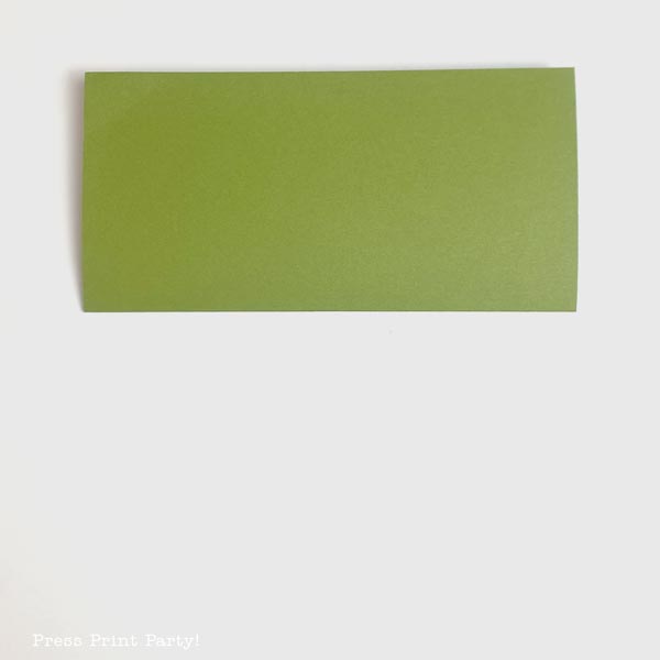 A green rectangle paper is positioned centrally on a light gray background. The smooth texture and even coloring evoke the calm of a folded Christmas tree. In the bottom left corner, there is faint text that is partially visible.